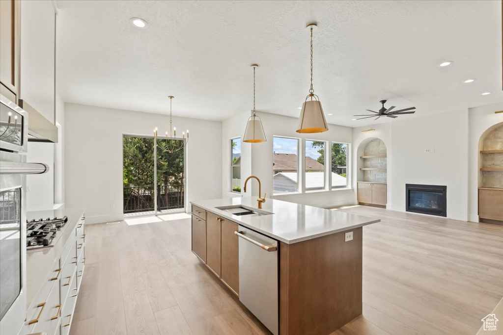 Kitchen featuring a glass covered fireplace, appliances with stainless steel finishes, a sink, light wood-style flooring, and an island with sink
