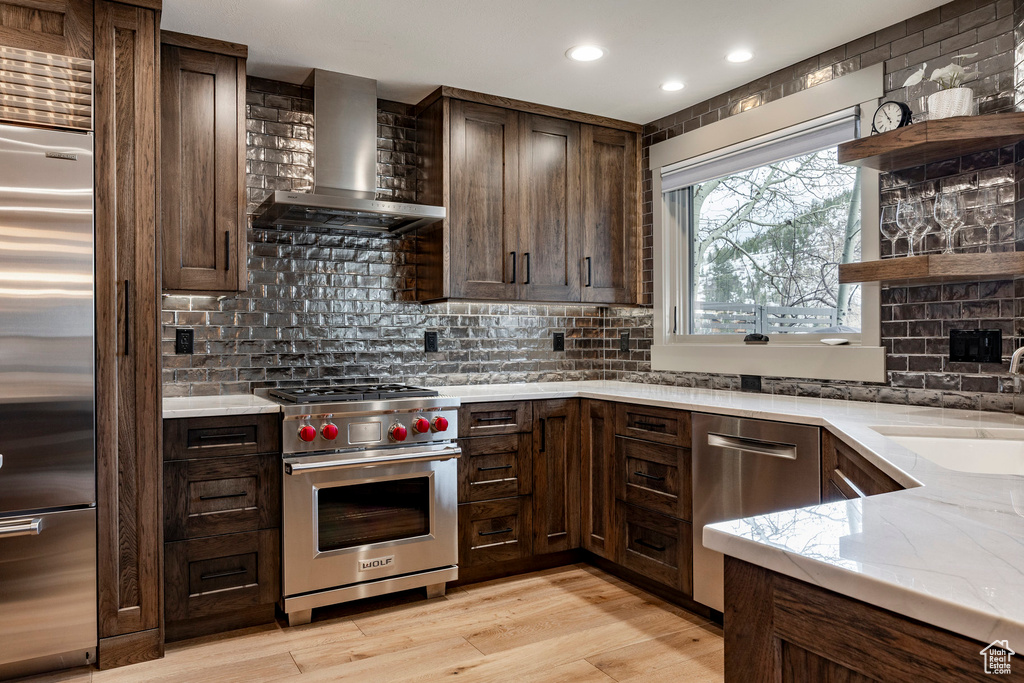 Kitchen featuring open shelves, light wood-style flooring, light stone countertops, premium appliances, and wall chimney exhaust hood