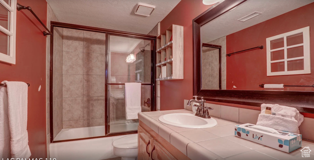 Full bath with a textured ceiling, visible vents, bath / shower combo with glass door, toilet, and vanity