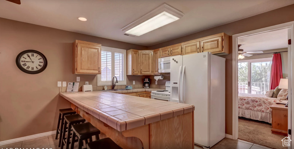 Kitchen featuring white appliances, light brown cabinetry, a breakfast bar, a sink, and a peninsula