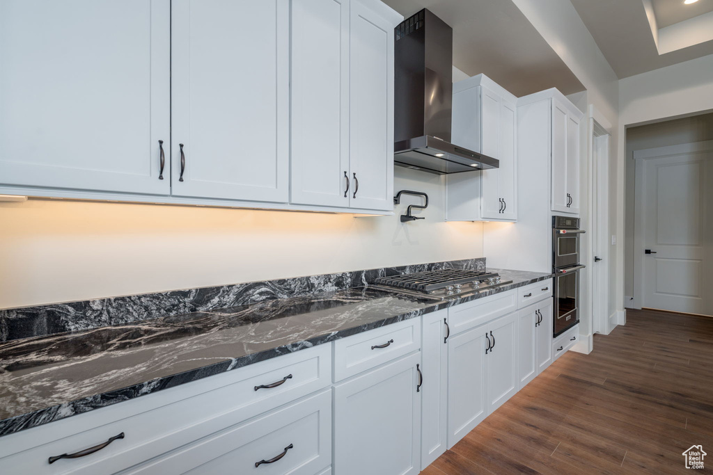 Kitchen featuring wall chimney exhaust hood, appliances with stainless steel finishes, white cabinets, dark stone counters, and dark wood-style flooring