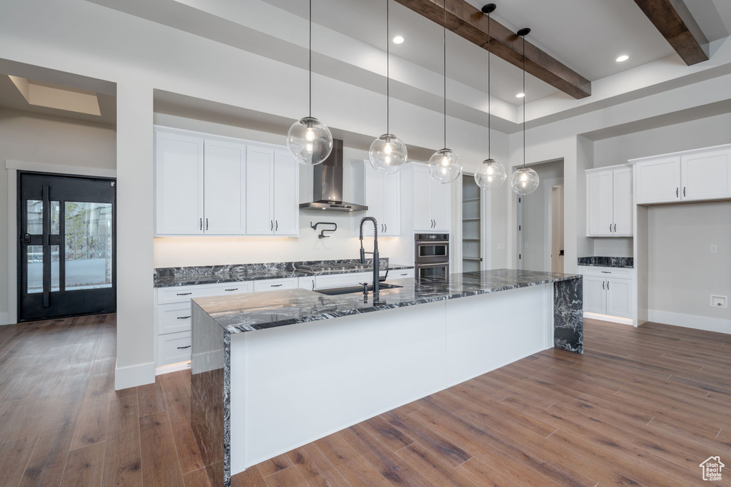 Kitchen featuring wall chimney exhaust hood, beam ceiling, dark stone counters, white cabinets, and a large island with sink