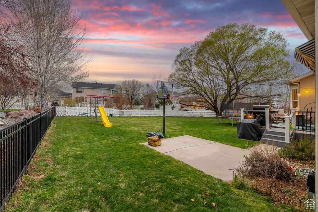 Yard at dusk with a wooden deck, a playground, a patio, a trampoline, and a fenced backyard