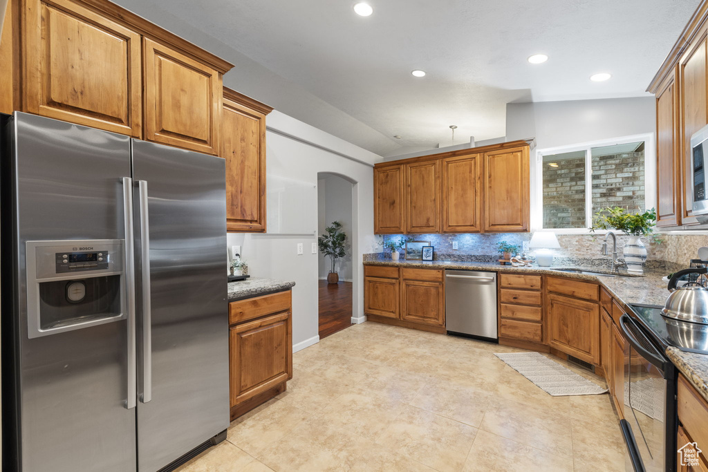 Kitchen featuring decorative backsplash, brown cabinets, stainless steel appliances, arched walkways, and stone counters