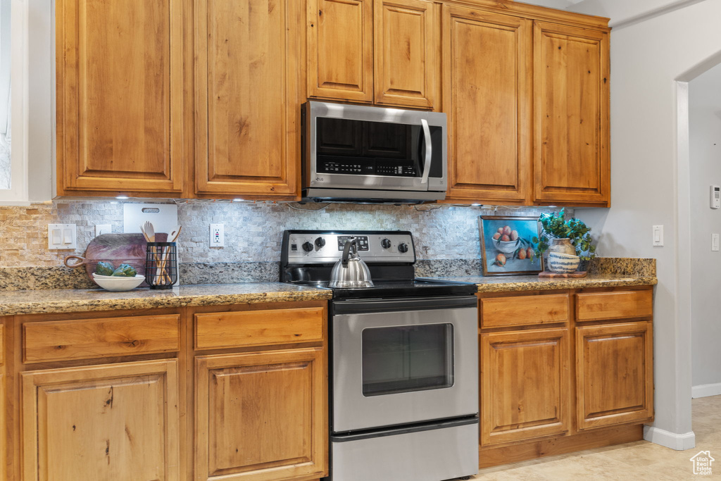 Kitchen with stainless steel appliances, baseboards, decorative backsplash, light stone countertops, and brown cabinets