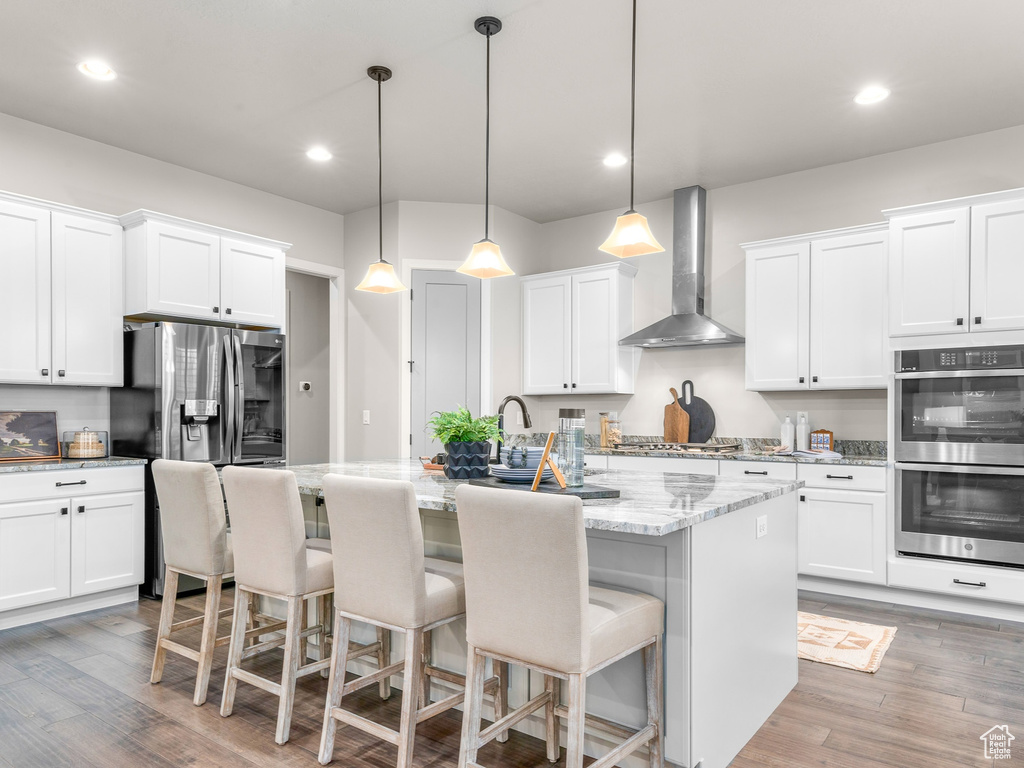 Kitchen featuring white cabinets, a center island with sink, appliances with stainless steel finishes, light wood-type flooring, and wall chimney range hood