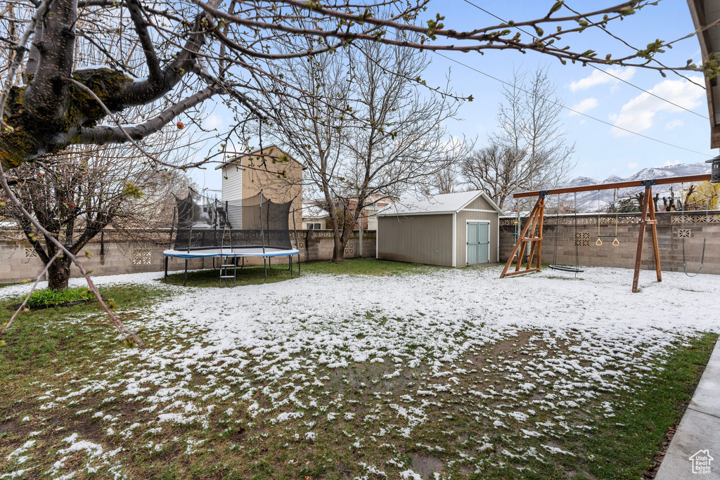 Snowy yard with a playground, a trampoline, a fenced backyard, a storage unit, and an outbuilding