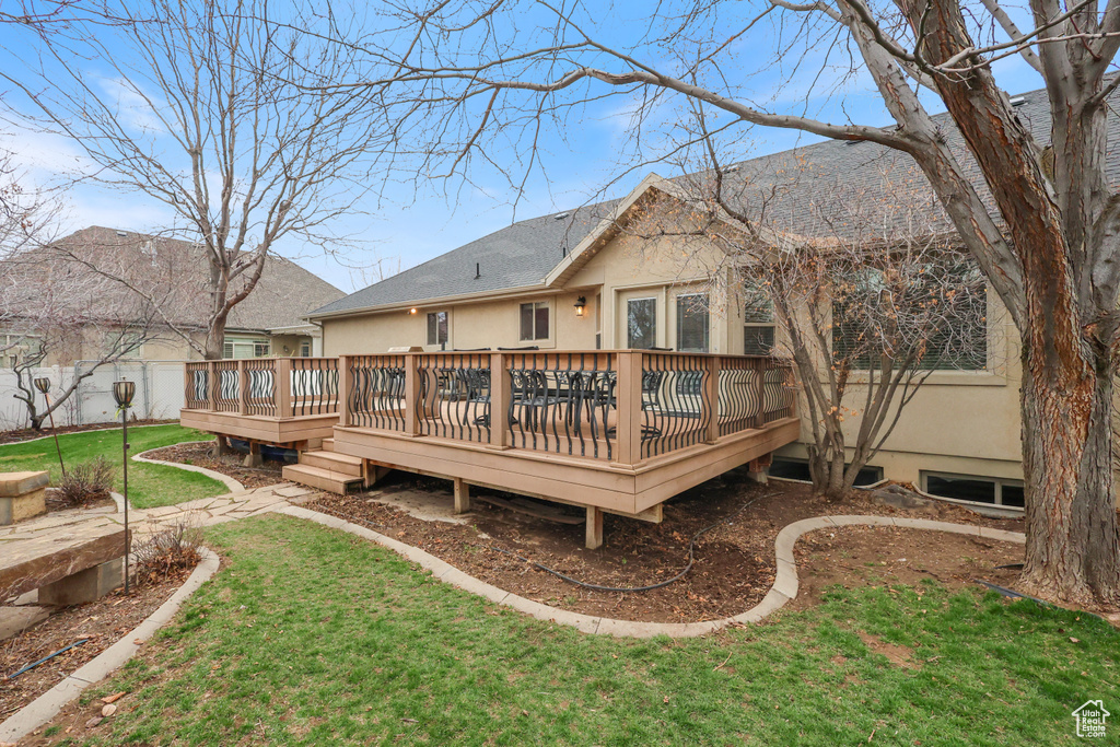 Rear view of house with stucco siding, fence, a wooden deck, roof with shingles, and a lawn