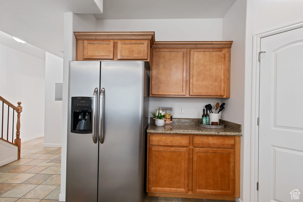 Kitchen with light tile patterned flooring, stainless steel fridge, brown cabinetry, dark stone countertops, and baseboards