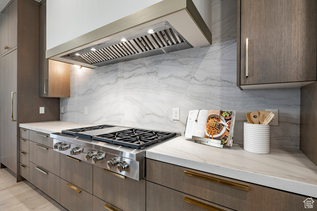 Kitchen featuring wall chimney range hood, stainless steel gas stovetop, backsplash, light stone countertops, and dark brown cabinets