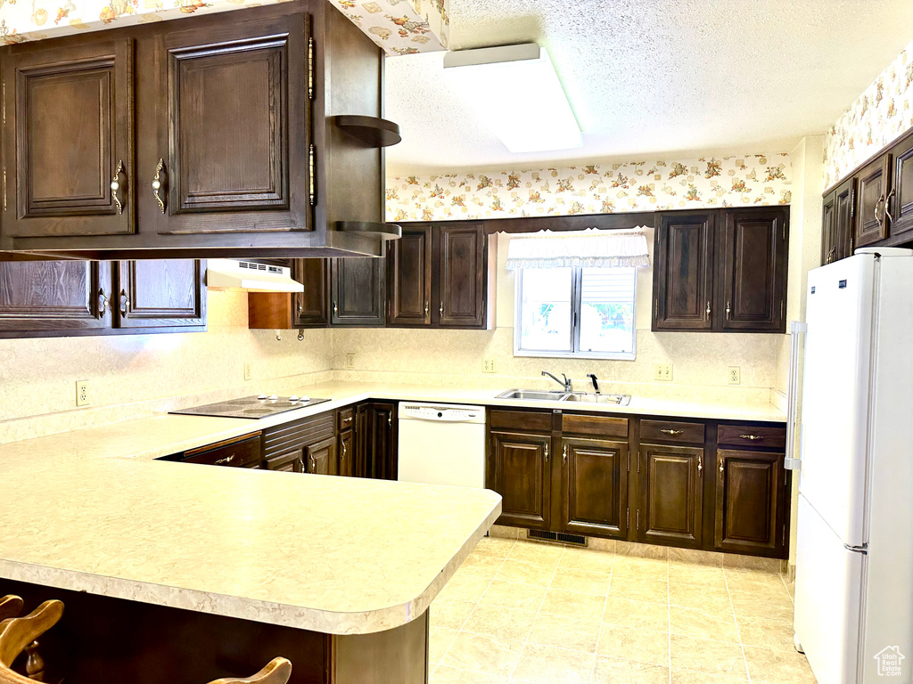 Kitchen featuring white appliances, light countertops, dark brown cabinetry, under cabinet range hood, and a textured ceiling