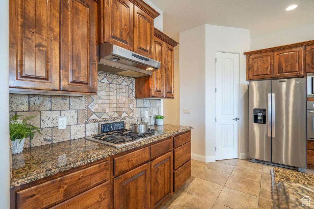 Kitchen featuring backsplash, appliances with stainless steel finishes, light tile patterned flooring, dark stone countertops, and under cabinet range hood