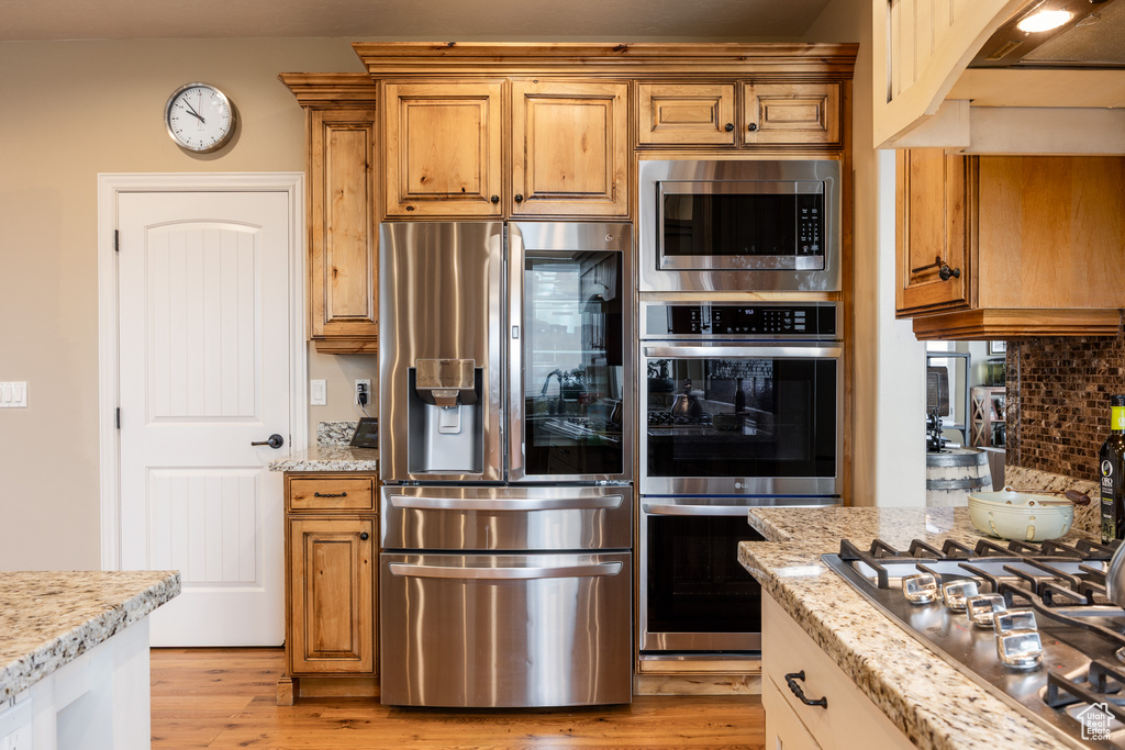 Kitchen featuring light stone counters, backsplash, appliances with stainless steel finishes, light wood-type flooring, and brown cabinetry