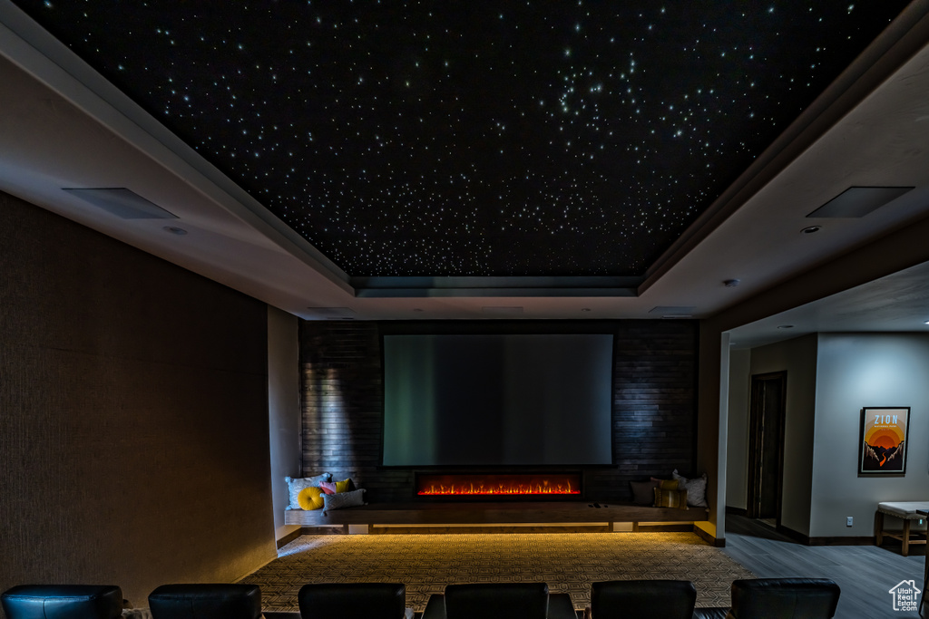Home theater room with a raised ceiling, wood finished floors, and a warm lit fireplace