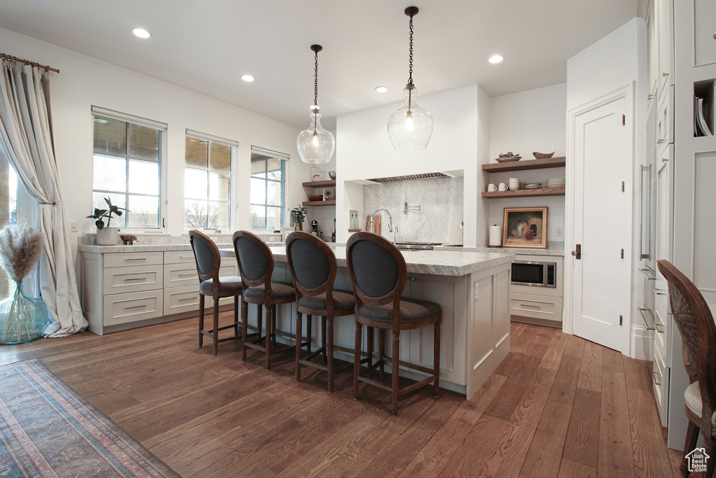 Kitchen featuring light countertops, a kitchen island with sink, stainless steel microwave, dark wood finished floors, and open shelves