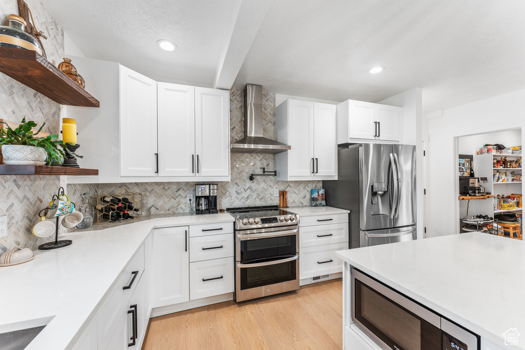 Kitchen with appliances with stainless steel finishes, white cabinetry, open shelves, light wood-style floors, and wall chimney range hood
