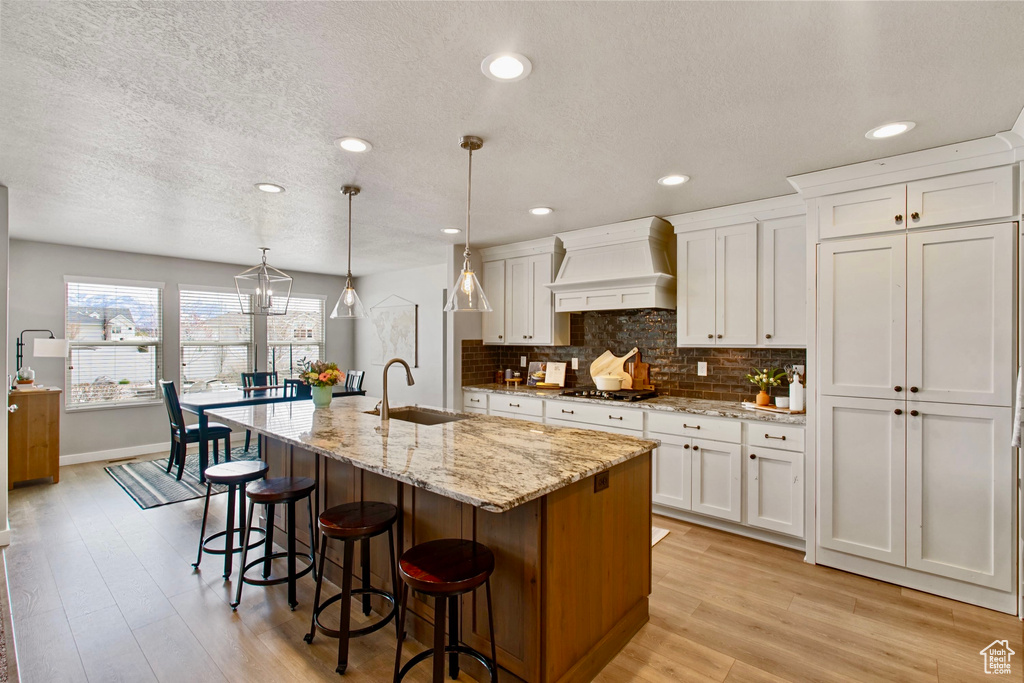 Kitchen with a sink, premium range hood, stainless steel gas stovetop, light wood-style floors, and decorative backsplash