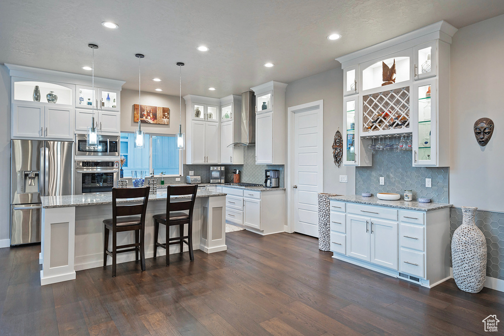 Kitchen with wall chimney exhaust hood, appliances with stainless steel finishes, white cabinetry, dark wood-style floors, and a center island