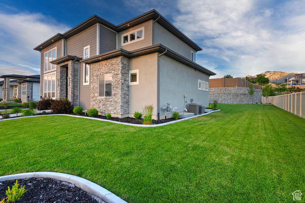 View of side of property featuring cooling unit, stucco siding, fence, stone siding, and a yard