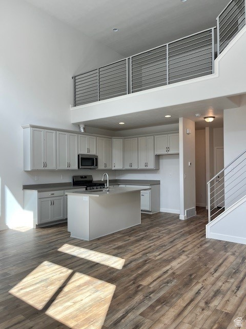 Kitchen featuring a towering ceiling, an island with sink, dark wood-style floors, baseboards, and appliances with stainless steel finishes