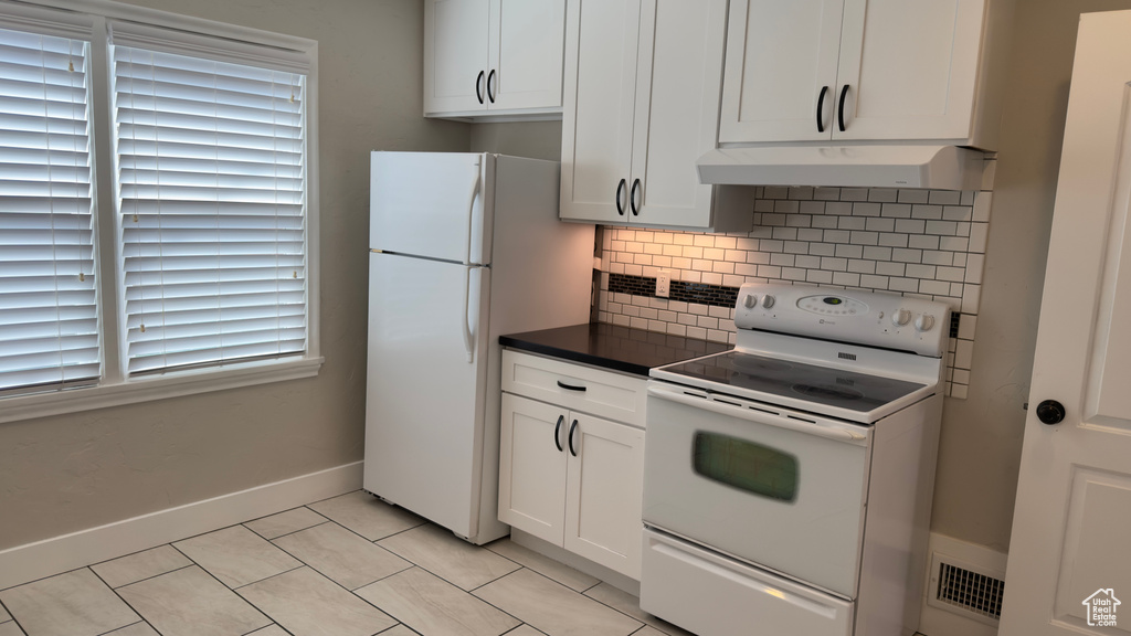 Kitchen with white cabinetry, white appliances, under cabinet range hood, backsplash, and dark countertops