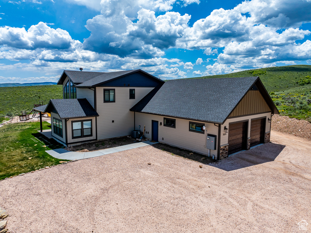 View of front facade featuring driveway, roof with shingles, stone siding, and central AC