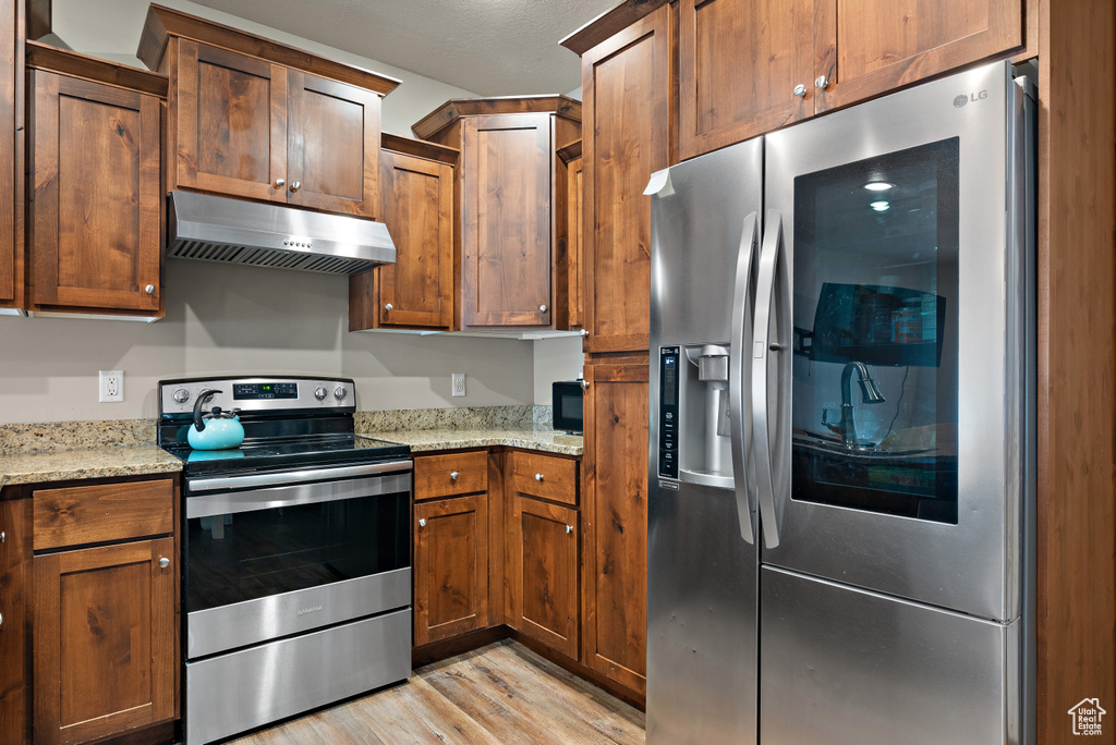 Kitchen with light stone countertops, light wood-type flooring, under cabinet range hood, stainless steel appliances, and brown cabinets