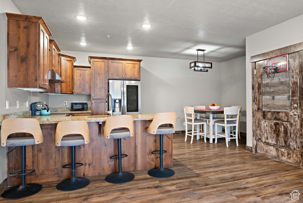 Kitchen featuring dark wood-style flooring, a peninsula, stainless steel refrigerator with ice dispenser, under cabinet range hood, and brown cabinetry