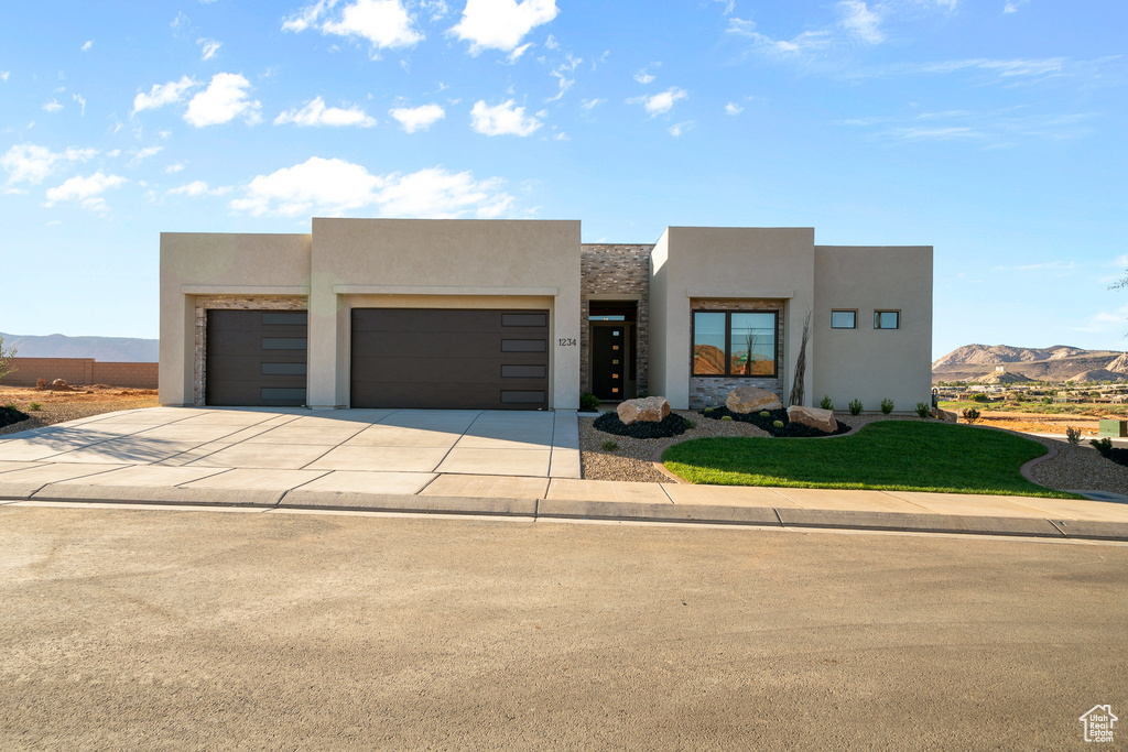 Pueblo-style house with stucco siding, an attached garage, concrete driveway, a front lawn, and a mountain view