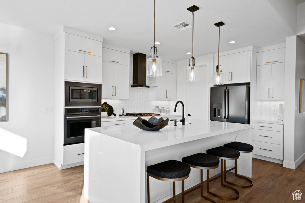 Kitchen featuring appliances with stainless steel finishes, wall chimney range hood, backsplash, a kitchen breakfast bar, and light wood finished floors