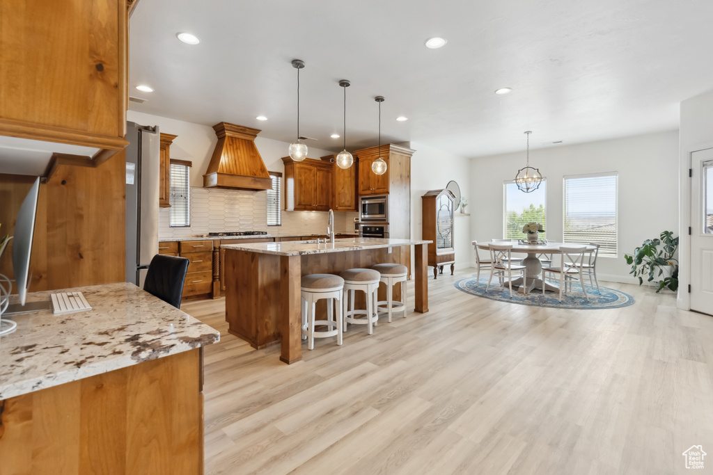 Kitchen with custom range hood, backsplash, light wood-style floors, brown cabinets, and appliances with stainless steel finishes