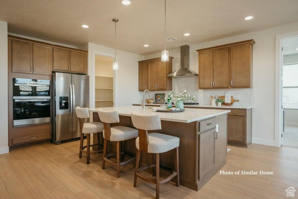 Kitchen with stainless steel appliances, light wood-style floors, a center island with sink, and wall chimney exhaust hood