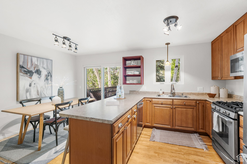 Kitchen featuring appliances with stainless steel finishes, light wood-style flooring, a peninsula, brown cabinetry, and decorative light fixtures