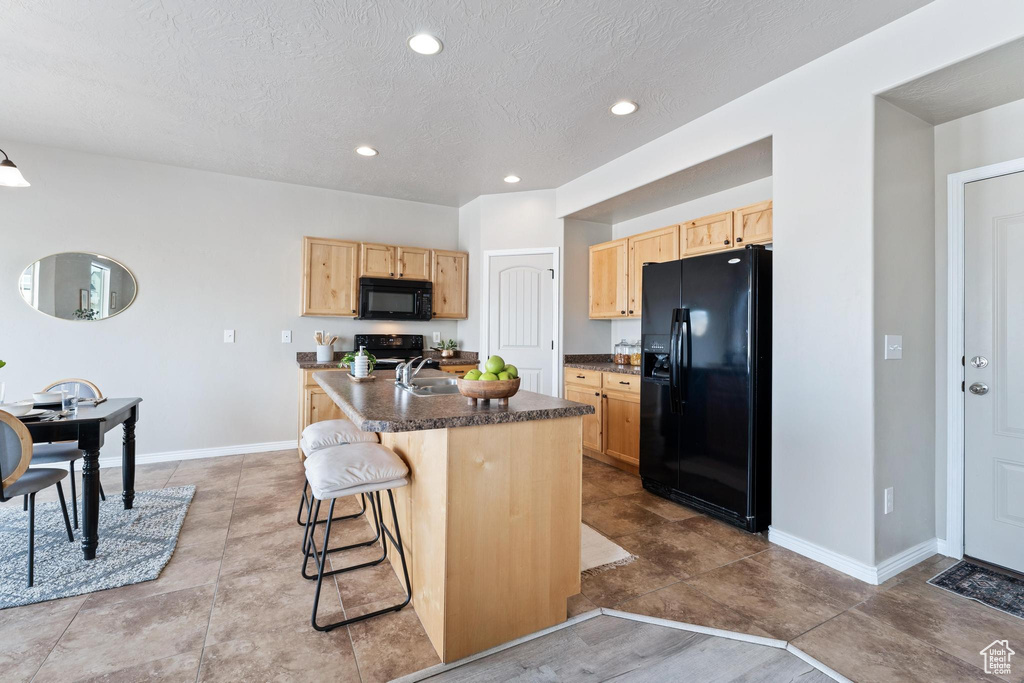 Kitchen featuring light brown cabinetry, black appliances, a breakfast bar, a sink, and dark countertops