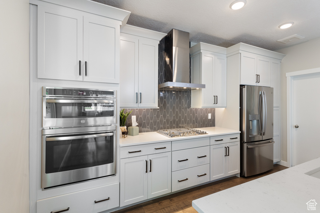 Kitchen featuring stainless steel appliances, wall chimney exhaust hood, tasteful backsplash, dark wood-style floors, and white cabinetry