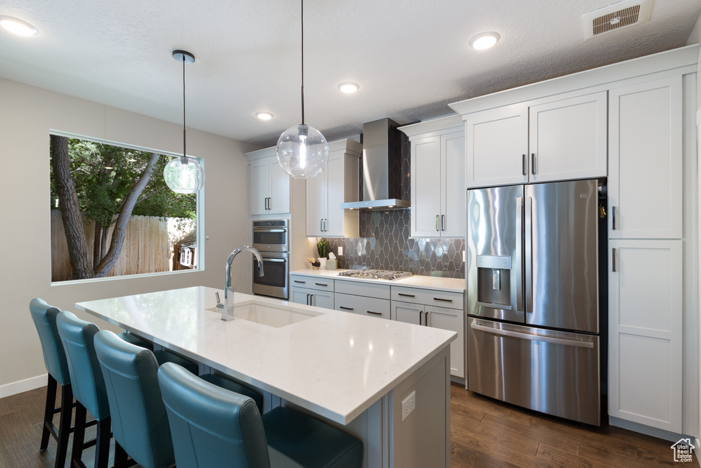 Kitchen with stainless steel appliances, wall chimney range hood, a sink, a center island with sink, and light countertops
