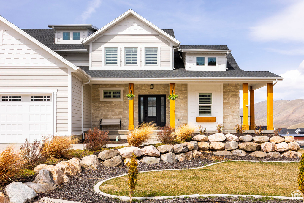 View of front facade with covered porch, a shingled roof, stone siding, a garage, and a mountain view