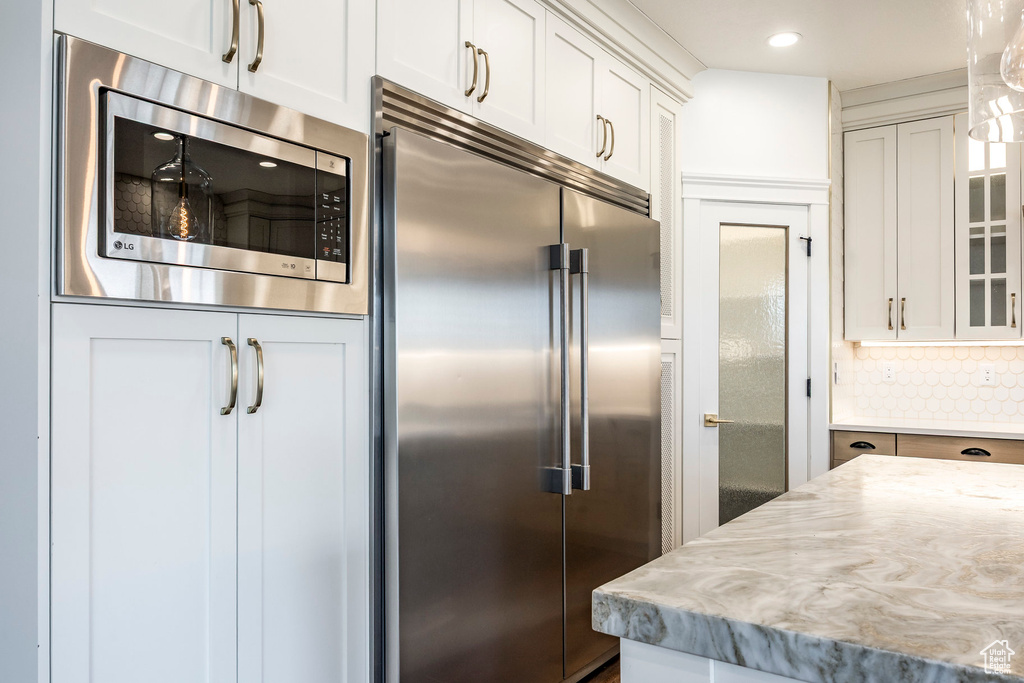 Kitchen featuring built in appliances, glass insert cabinets, backsplash, white cabinetry, and light stone countertops