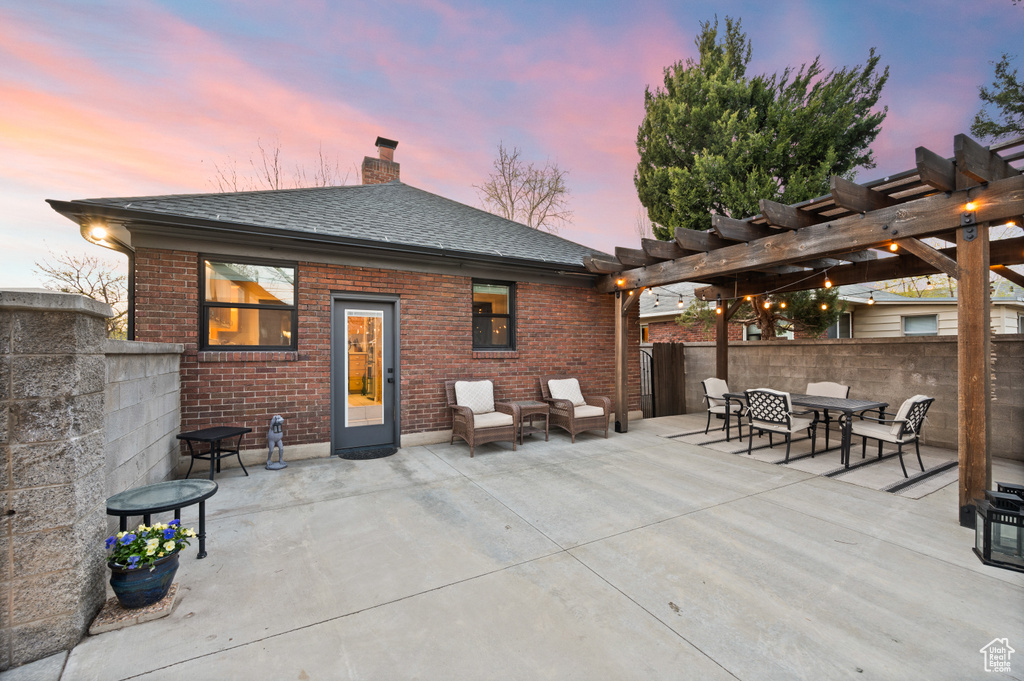 Back of house at dusk featuring a pergola, a patio area, fence, brick siding, and a chimney