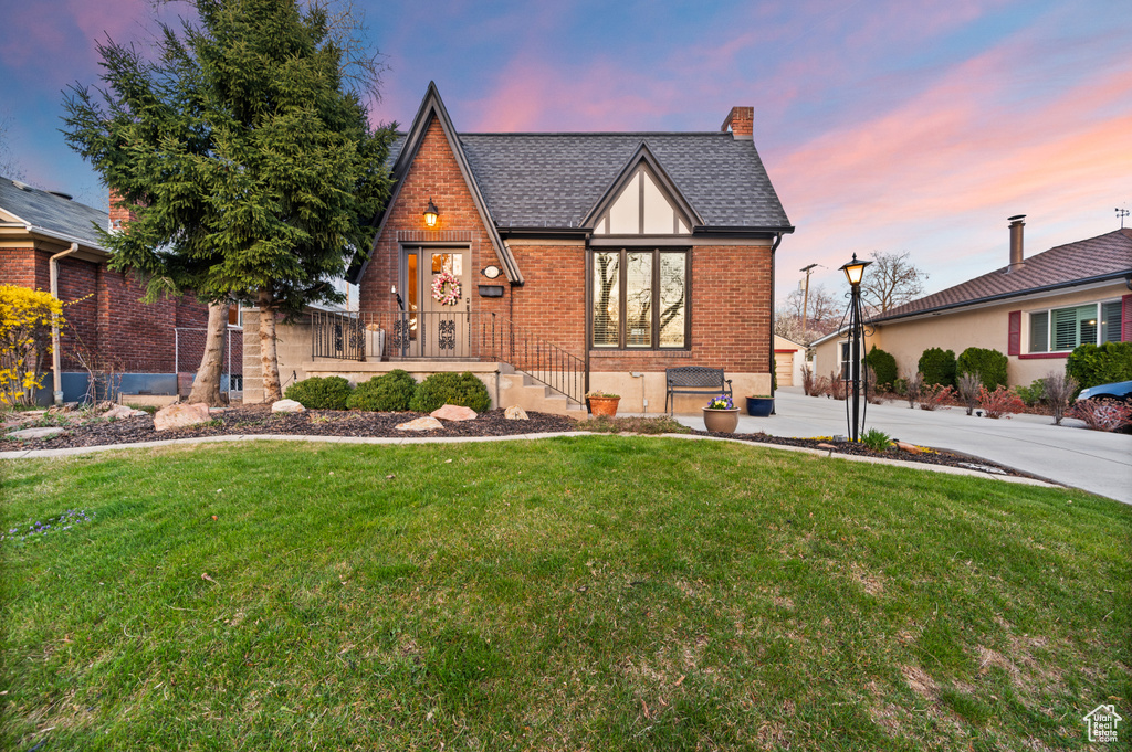 View of front of property with a front yard, a shingled roof, a chimney, concrete driveway, and brick siding