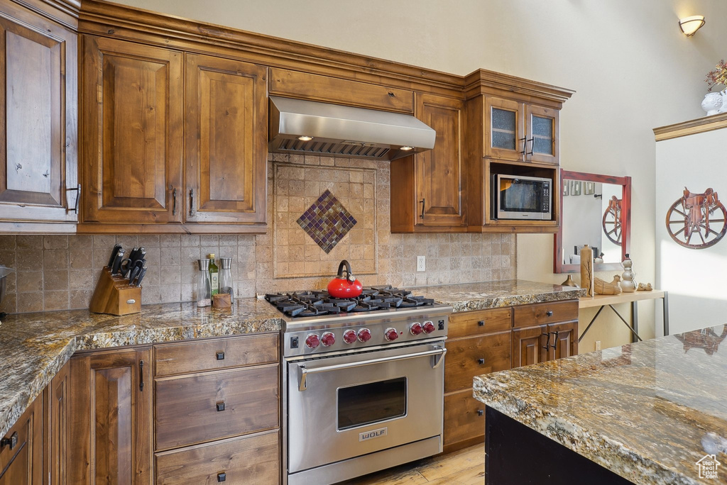 Kitchen with range hood, glass insert cabinets, tasteful backsplash, appliances with stainless steel finishes, and brown cabinets