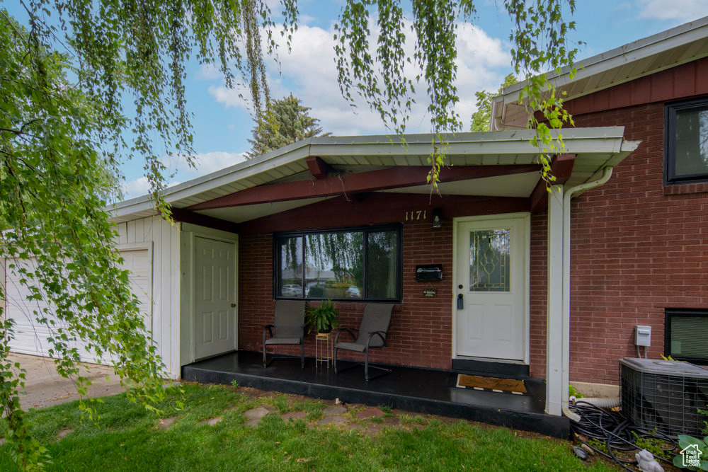 View of exterior entry with brick siding, a porch, central AC unit, and an attached garage