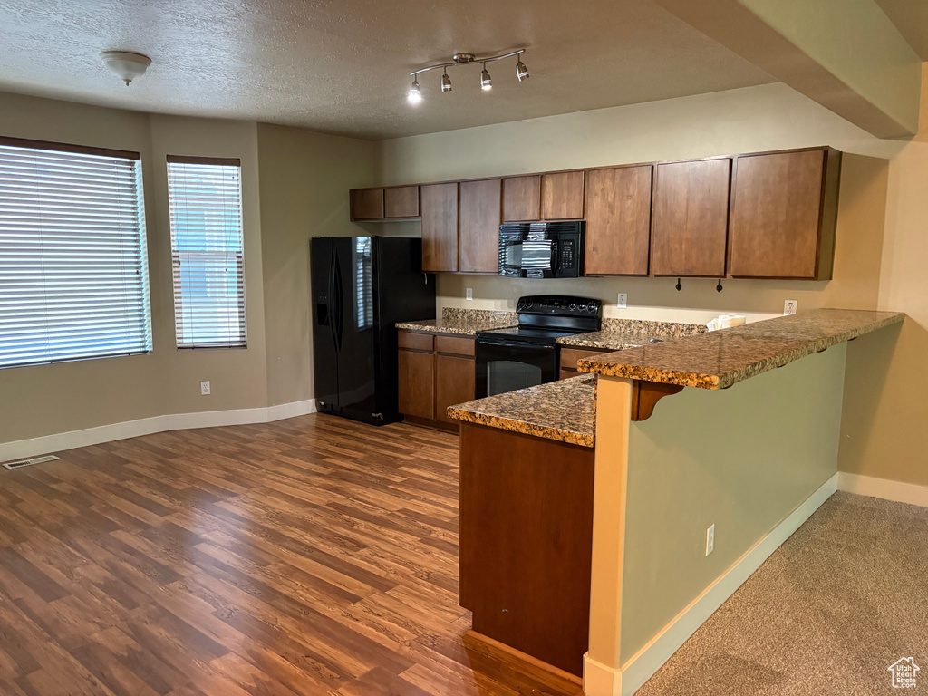 Kitchen with black appliances, a peninsula, dark wood-type flooring, brown cabinetry, and a kitchen bar