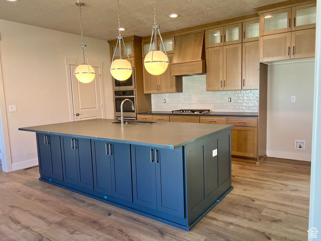Kitchen featuring custom range hood, appliances with stainless steel finishes, light wood-style flooring, decorative backsplash, and a kitchen island with sink