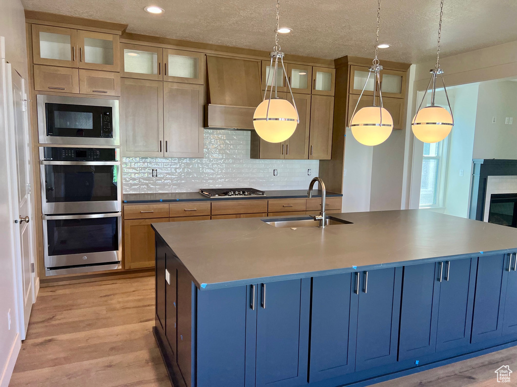 Kitchen featuring appliances with stainless steel finishes, light wood-style floors, custom exhaust hood, a center island with sink, and tasteful backsplash