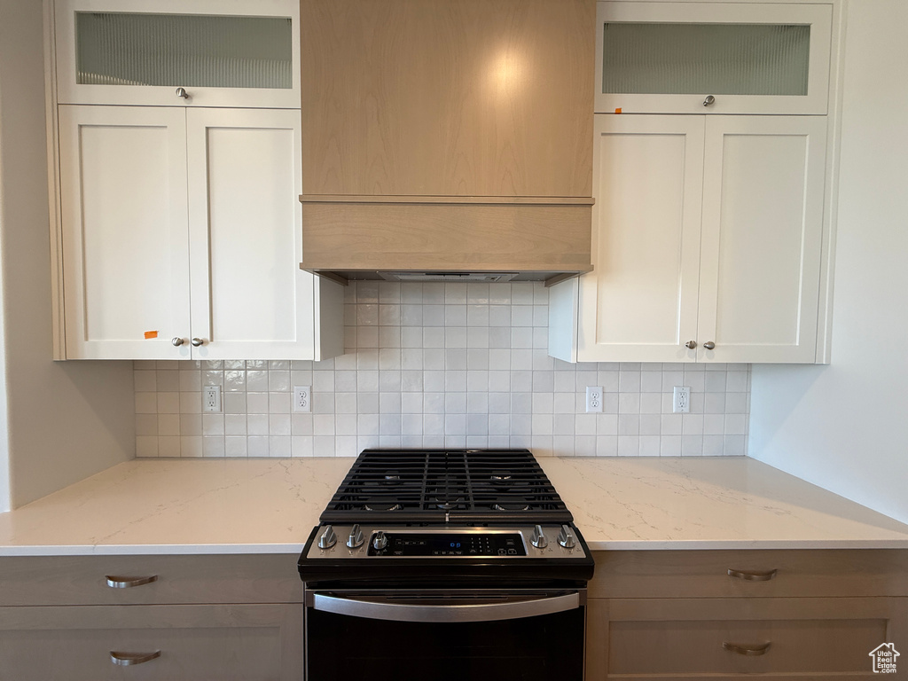 Kitchen featuring stainless steel gas stove, wall chimney range hood, tasteful backsplash, glass insert cabinets, and light stone counters