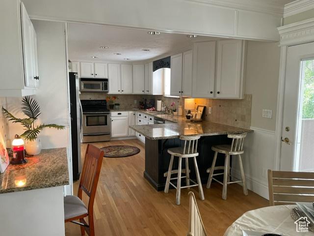 Kitchen featuring stainless steel appliances, a peninsula, light wood finished floors, white cabinetry, and stone counters