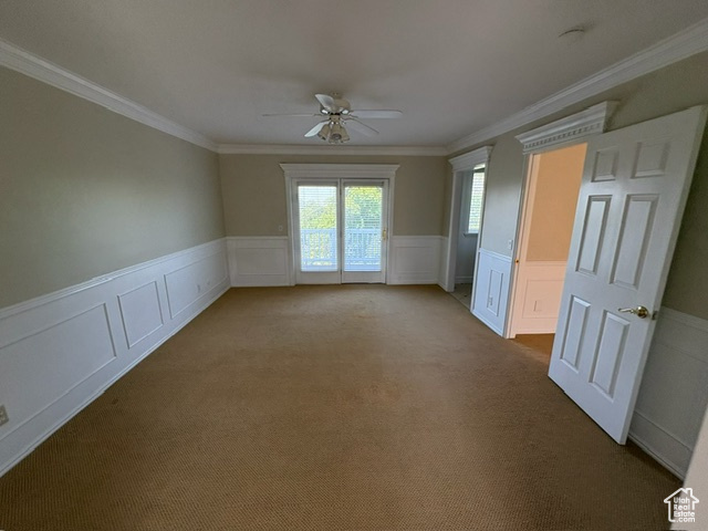 Carpeted empty room featuring ceiling fan, wainscoting, a decorative wall, and crown molding