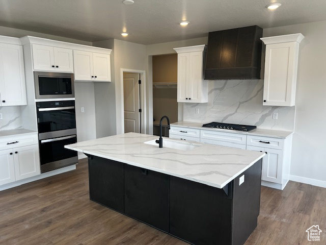 Kitchen featuring multiple ovens, custom exhaust hood, stainless steel microwave, a sink, and black cooktop