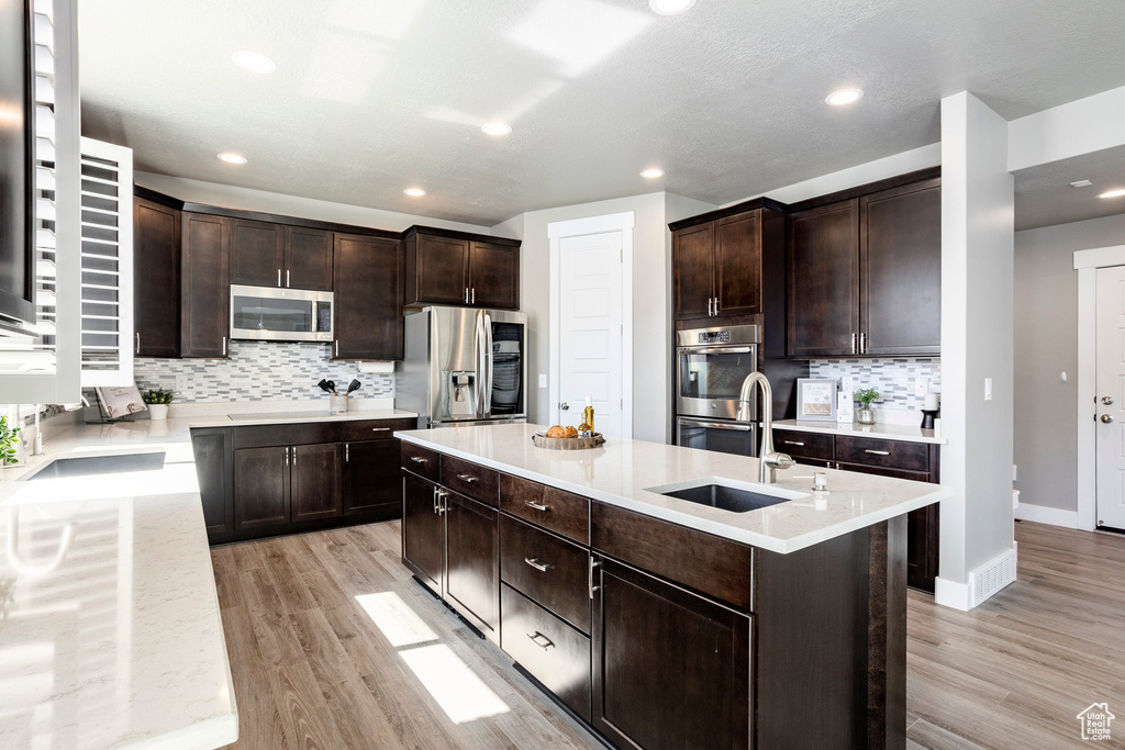 Kitchen featuring tasteful backsplash, dark brown cabinetry, an island with sink, light wood-style flooring, and appliances with stainless steel finishes